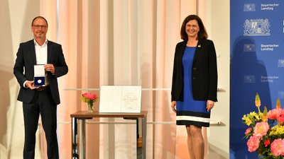 Robert Wagner präsentiert die soeben erhaltene Medaille. Daneben Landtagspräsidentin Ilse Aigner vor dem Logo des Bayerischen Landtages.