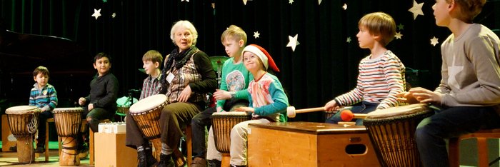 Kinder und Lehrerin spielen auf verschiedenen afrikanischen Trommeln, Weihnachtdeko im Hintergrund