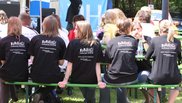 Menschen sitzen auf einer Bank, die Kamera zeigt ihre Rücken, sie tragen alle das gleiche schwarze T-Shirt mit dem Logo der Musikschule